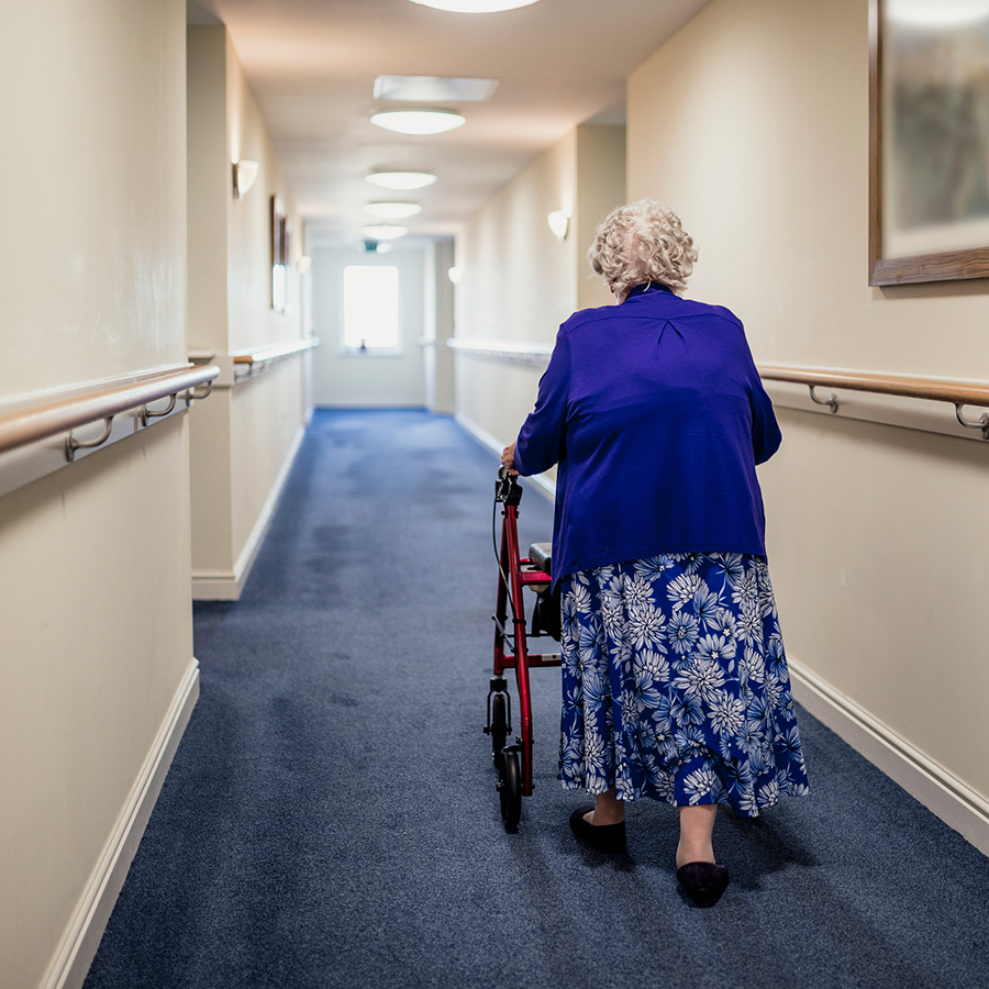 Une dame âgée circule avec sa marchette dans un corridor.