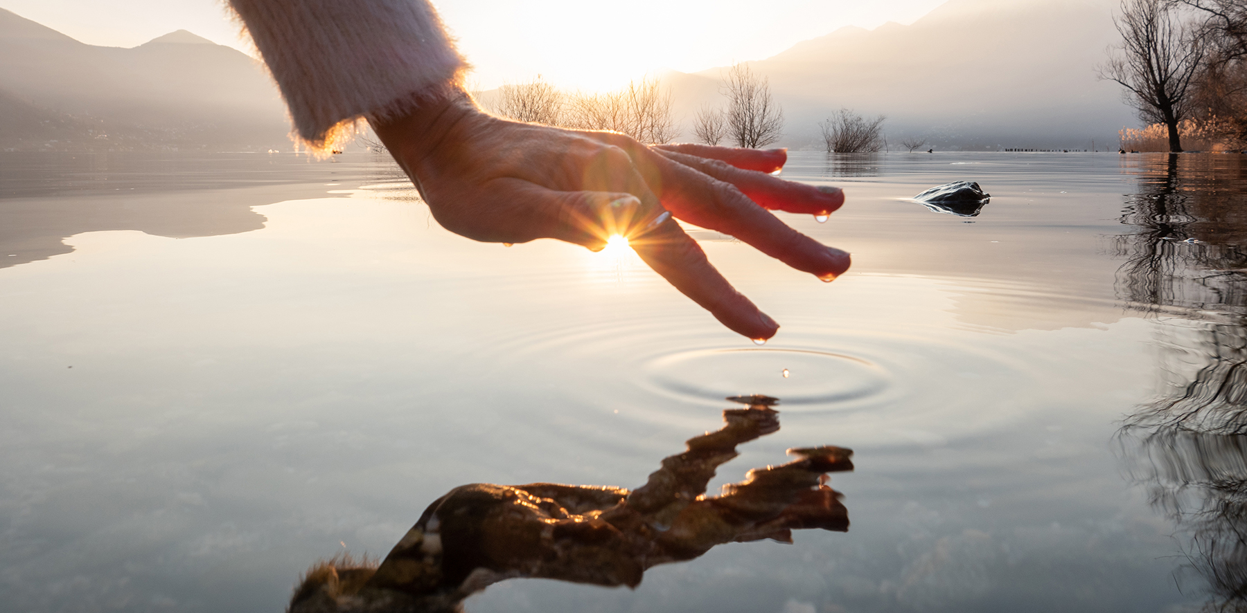 Une main laissant passer des rayons de soleil touche délicatement l’eau du lac