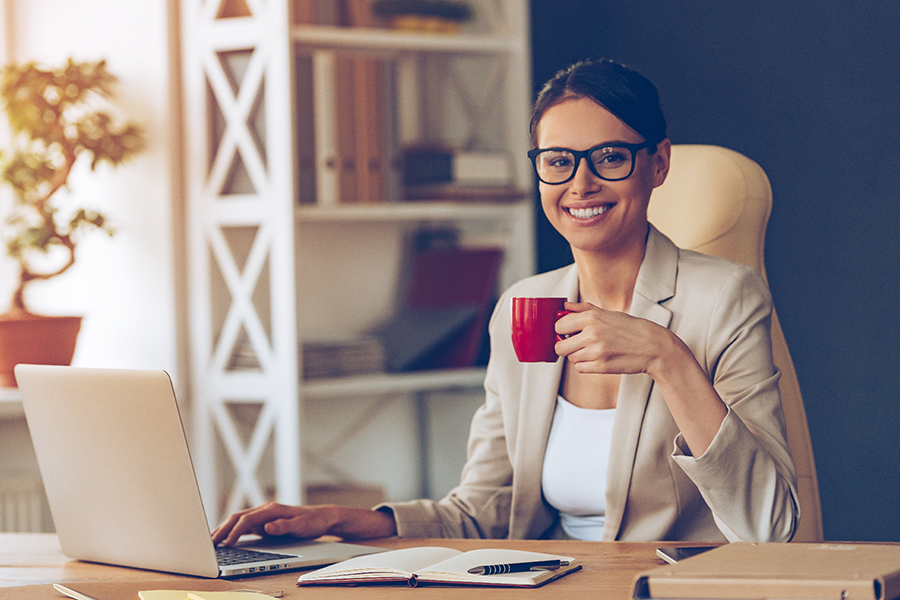 Une professionnel de la santé souriante est assise à son bureau avec une tasse de café dans une main et l’autre sur son portable