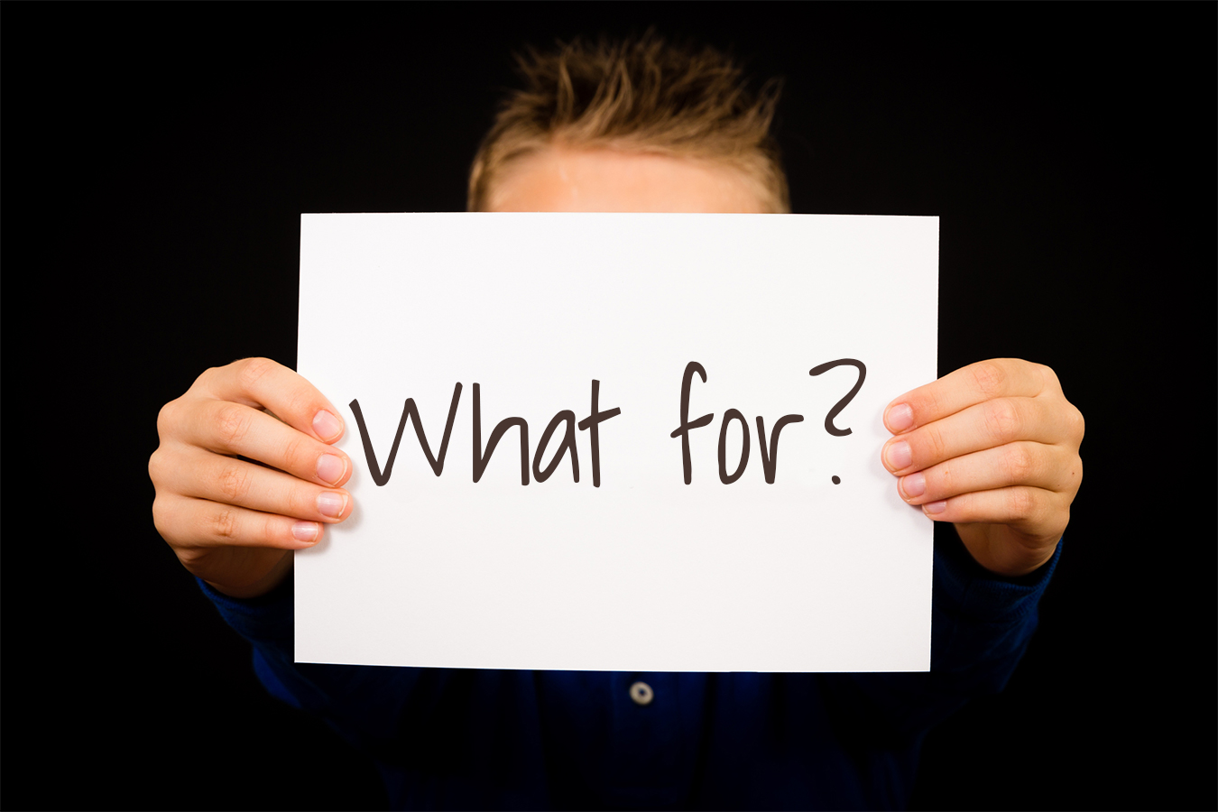 Hands holding a sheet displaying “WHAT” and “FOR” followed by a question mark. 